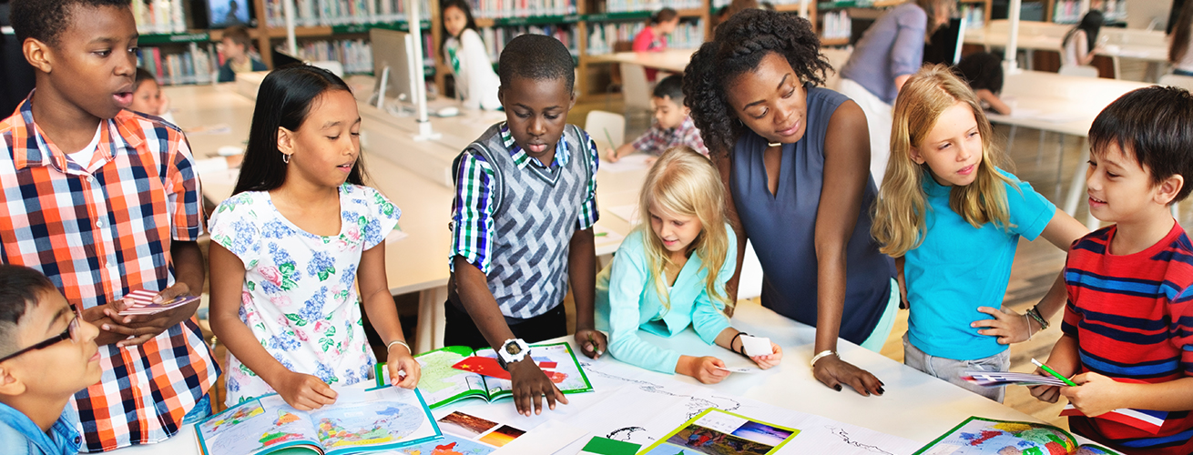 Teacher with students standing around a table looking at books and maps.